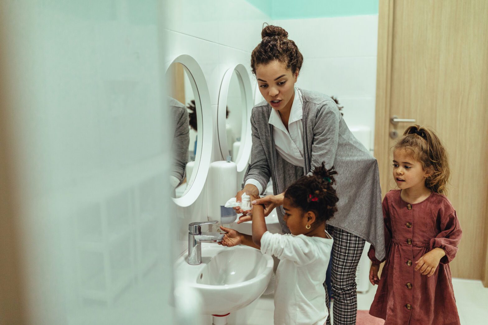 A teacher helping her students wash their hands in the school bathroom, practicing good hand hygiene.